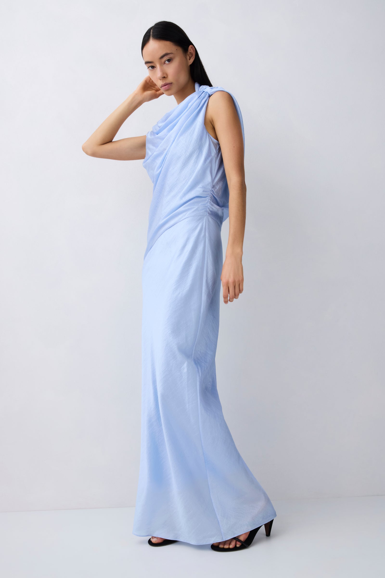 A woman modeling a light blue draped sleeveless maxi dress with a high neckline, standing against a plain light gray studio background.