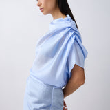 A model in profile wearing a light blue draped top and matching light blue skirt against a plain light gray studio background.
