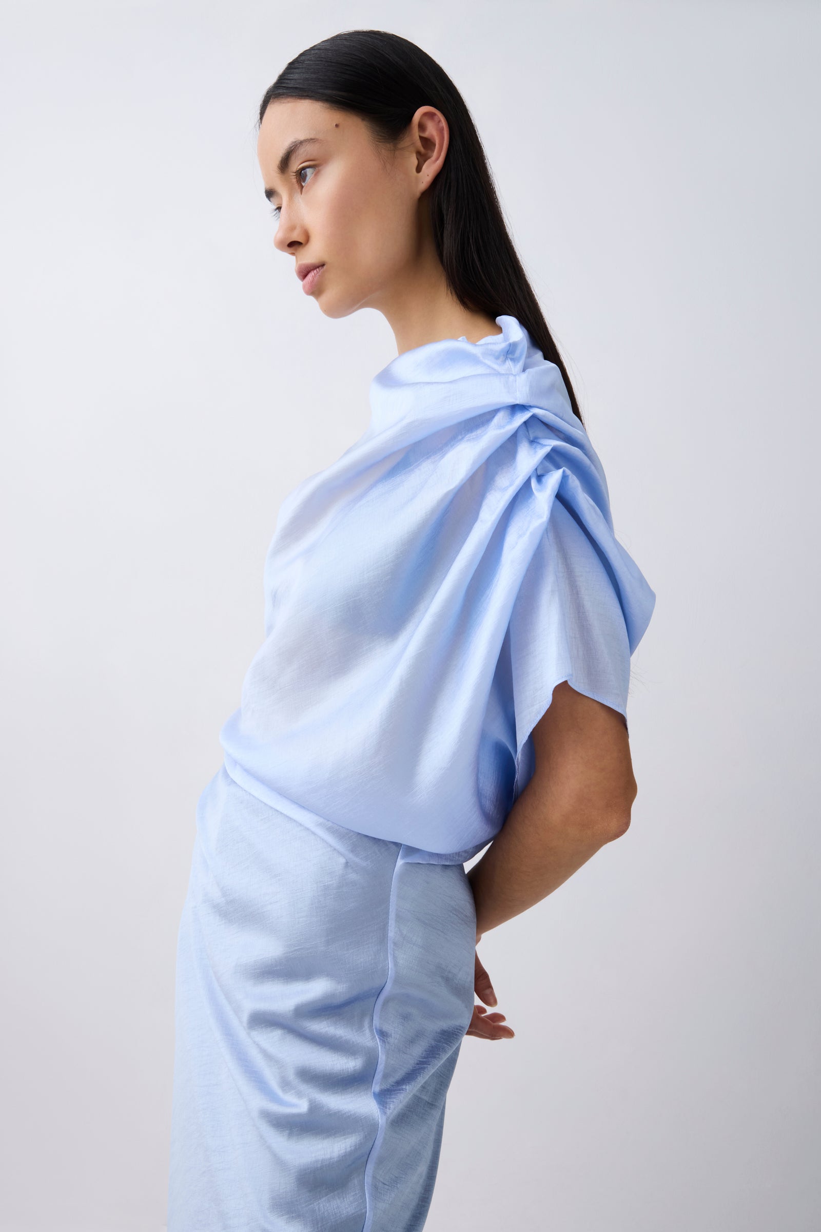 A model in profile wearing a light blue draped top and matching light blue skirt against a plain light gray studio background.