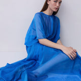 A woman wearing a flowing, semi-sheer royal blue dress sits against a plain light gray wall, holding the fabric of the dress with one hand.