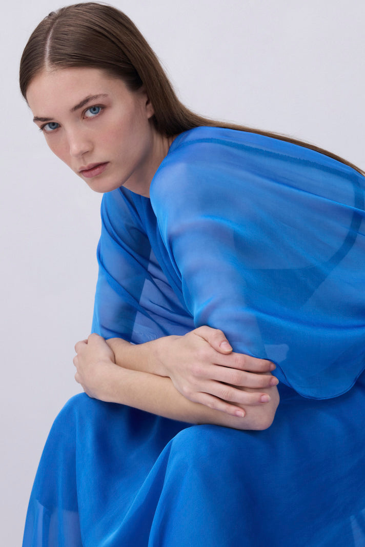 A woman wearing a flowing sheer royal blue dress or shawl, posed against a plain light gray studio background.