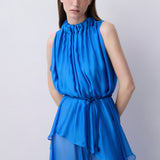 A woman modeling a sleeveless high-neck blue dress with a gathered peplum waist and a matching tie belt, photographed against a plain light gray studio background.