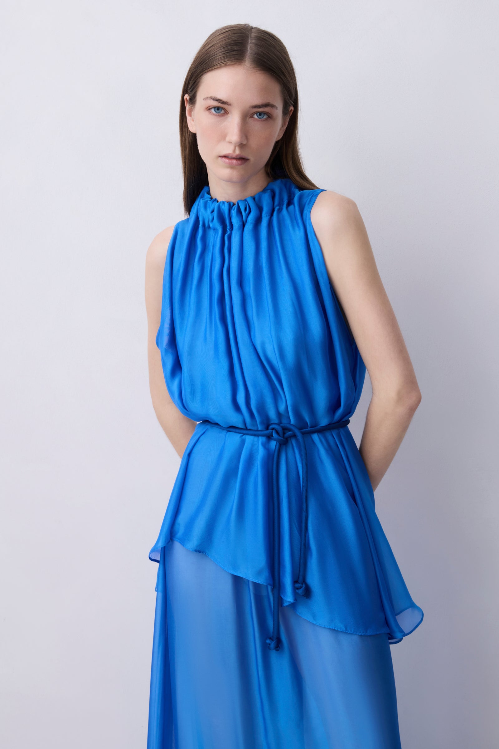 A woman modeling a sleeveless high-neck blue dress with a gathered peplum waist and a matching tie belt, photographed against a plain light gray studio background.