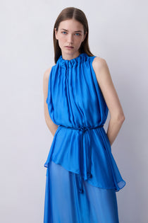 A woman modeling a sleeveless high-neck blue dress with a gathered peplum waist and a matching tie belt, photographed against a plain light gray studio background.