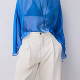A model wearing a sheer, semi-transparent royal blue long-sleeve top paired with off-white high-waisted trousers and a long black cord necklace with a clear heart-shaped pendant.