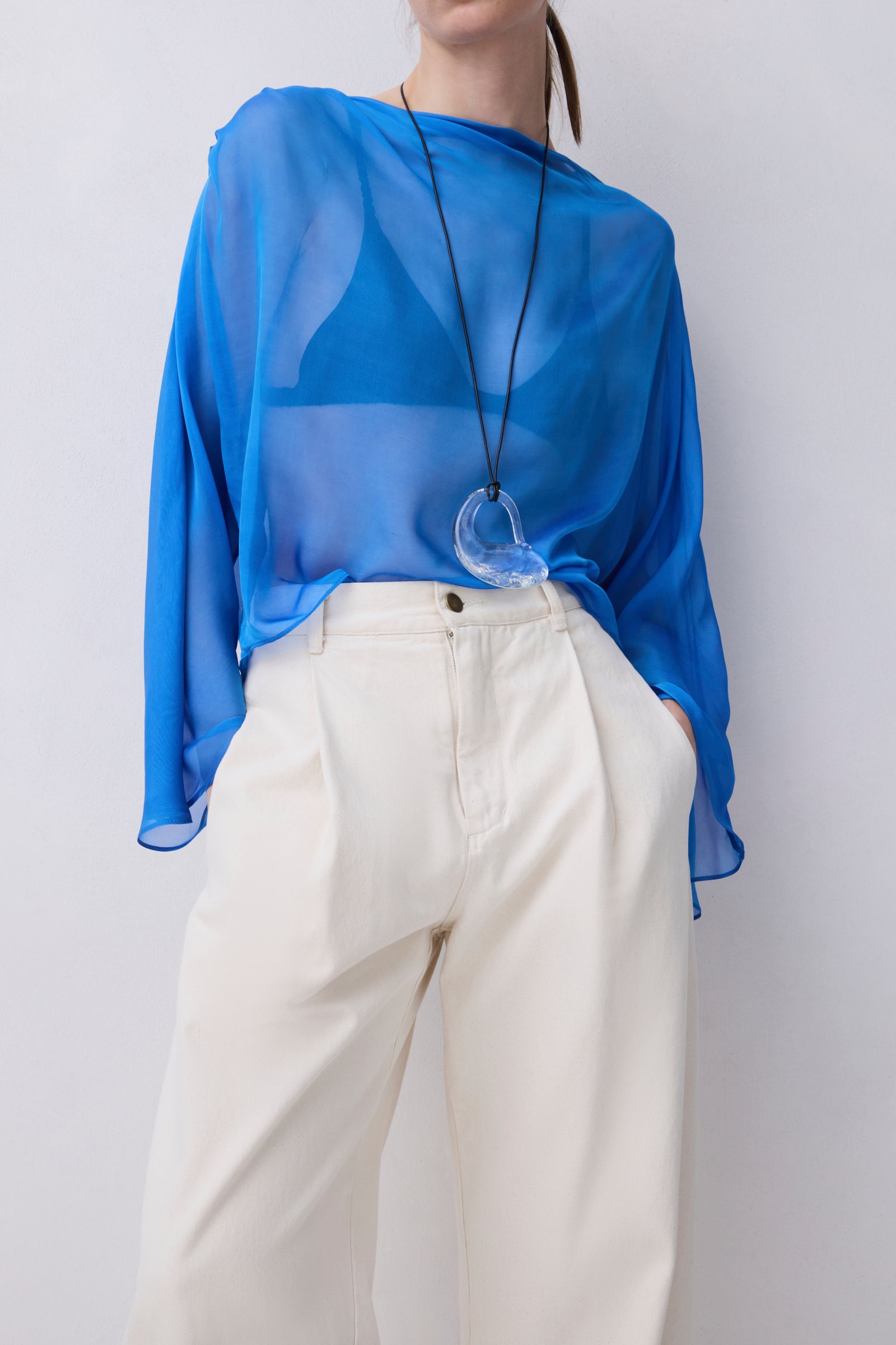 A model wearing a sheer, semi-transparent royal blue long-sleeve top paired with off-white high-waisted trousers and a long black cord necklace with a clear heart-shaped pendant.