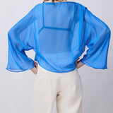 A woman viewed from behind wearing a sheer, semi-transparent blue long-sleeve blouse and cream-colored trousers, standing against a light gray studio background.