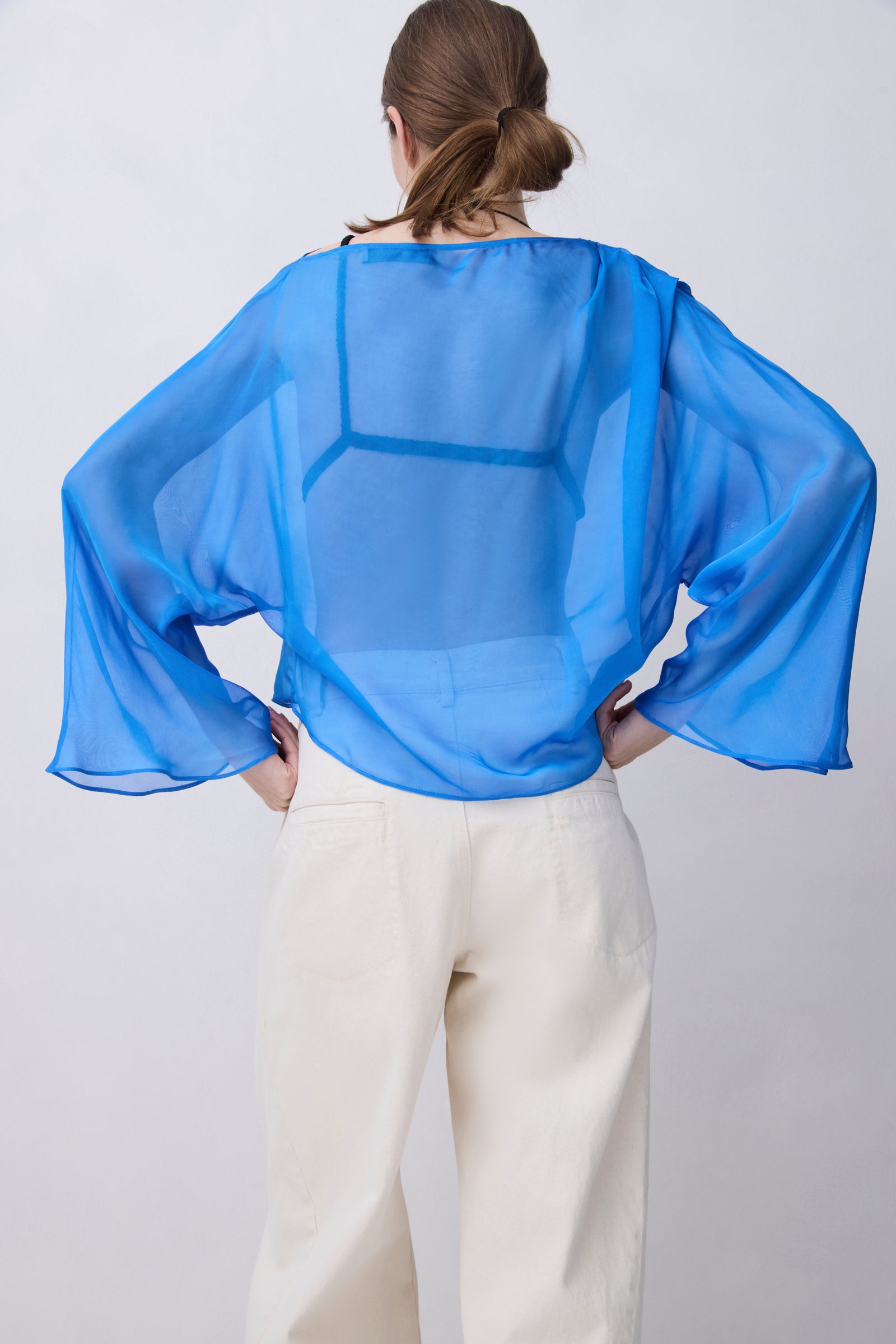A woman viewed from behind wearing a sheer, semi-transparent blue long-sleeve blouse and cream-colored trousers, standing against a light gray studio background.