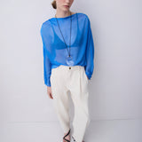 A woman modeling a sheer bright blue long-sleeve top with a wide neckline, paired with cream-colored wide-leg trousers and black sandals, standing against a plain light gray studio background.