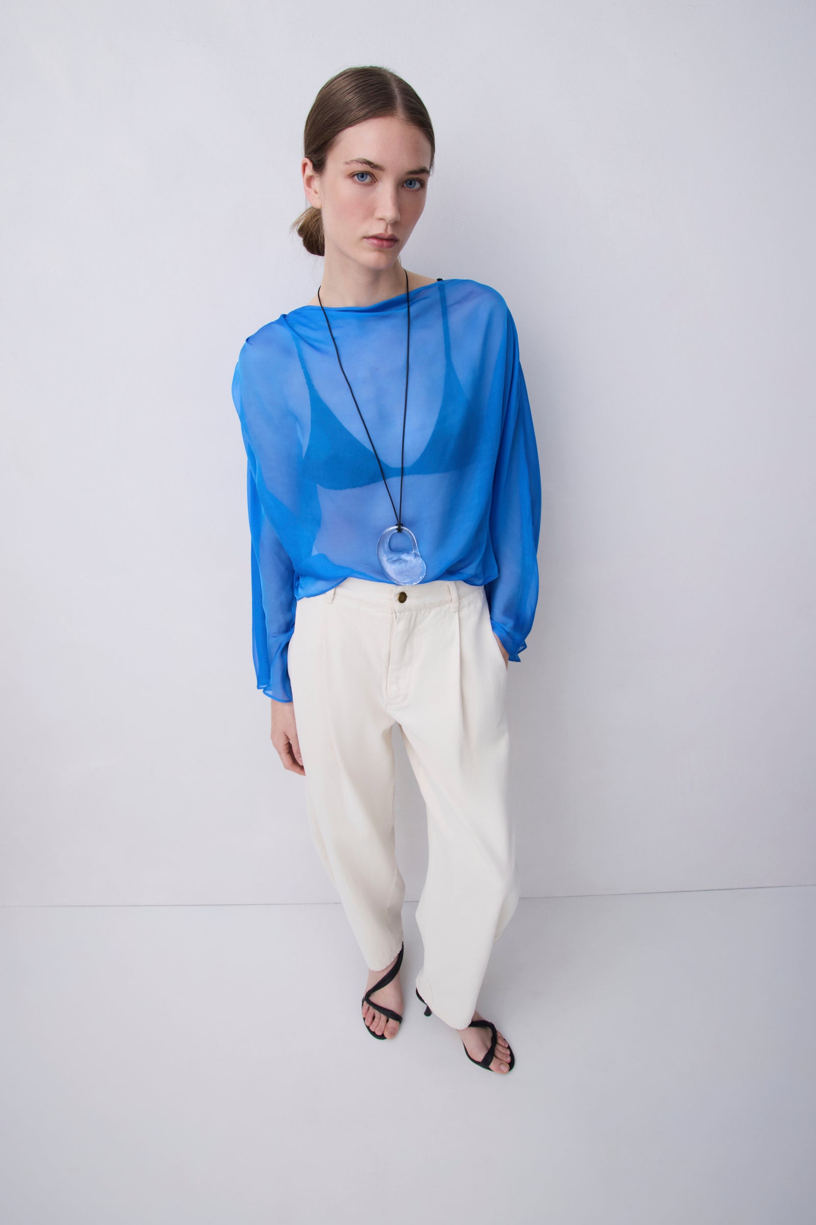 A woman modeling a sheer bright blue long-sleeve top with a wide neckline, paired with cream-colored wide-leg trousers and black sandals, standing against a plain light gray studio background.