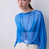 A woman wearing a sheer, long-sleeved blue top and cream-colored trousers, accessorized with a long black cord necklace featuring a large oval pendant, set against a plain light gray background.