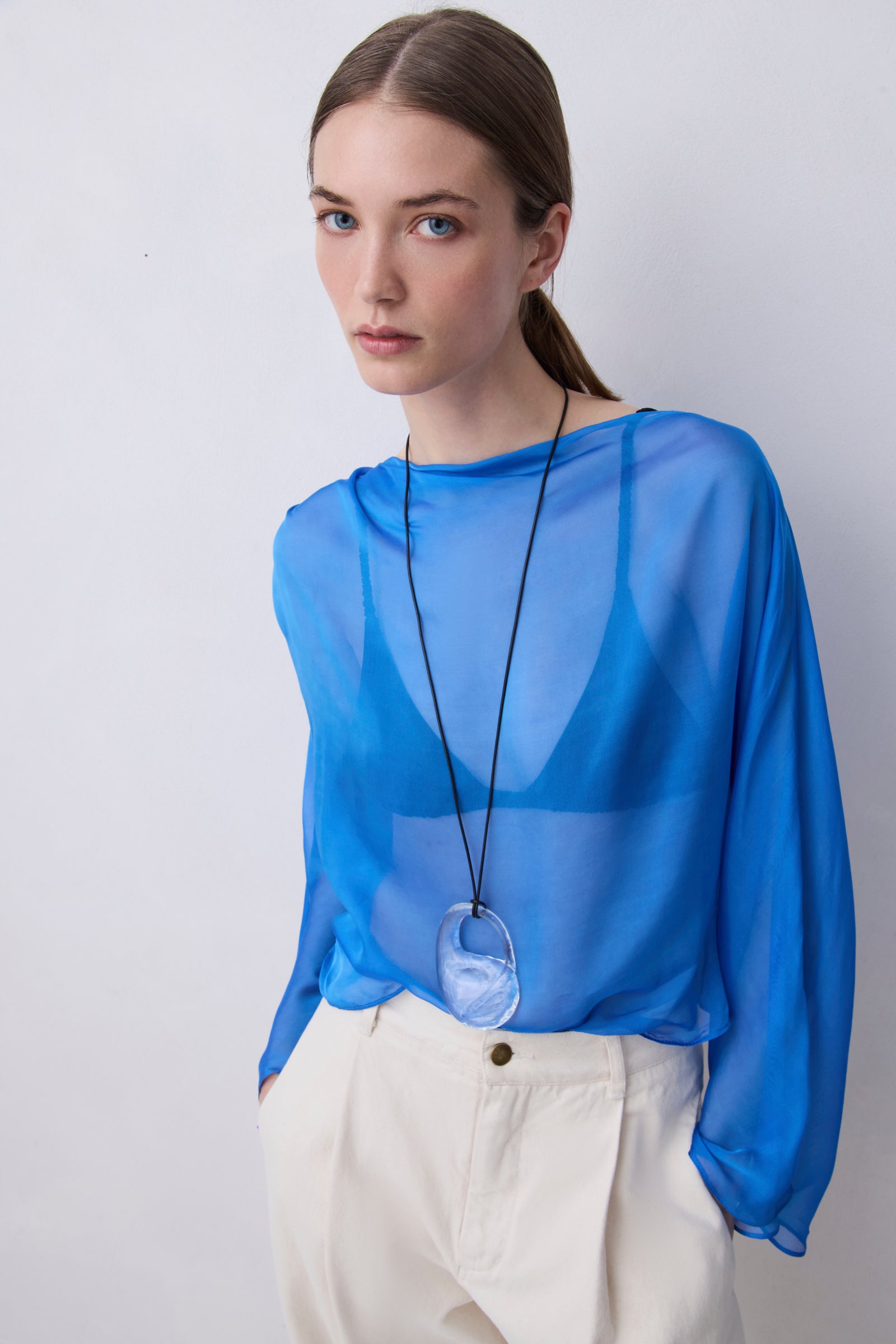A woman wearing a sheer, long-sleeved blue top and cream-colored trousers, accessorized with a long black cord necklace featuring a large oval pendant, set against a plain light gray background.