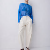 A model stands against a light gray studio wall wearing a sheer bright blue long-sleeve top, cream wide-leg trousers, and black strappy high-heeled sandals.