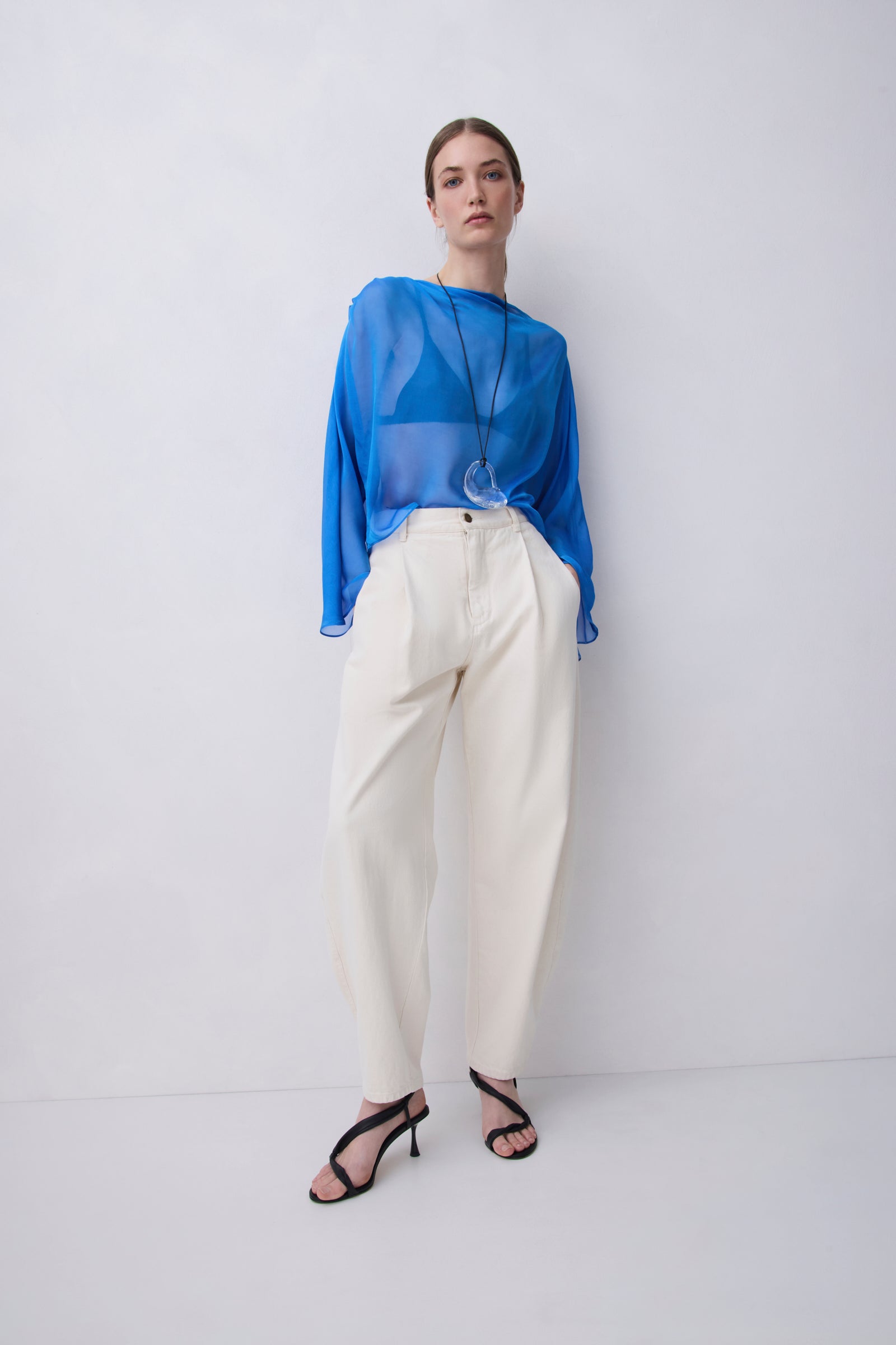 A model stands against a light gray studio wall wearing a sheer bright blue long-sleeve top, cream wide-leg trousers, and black strappy high-heeled sandals.