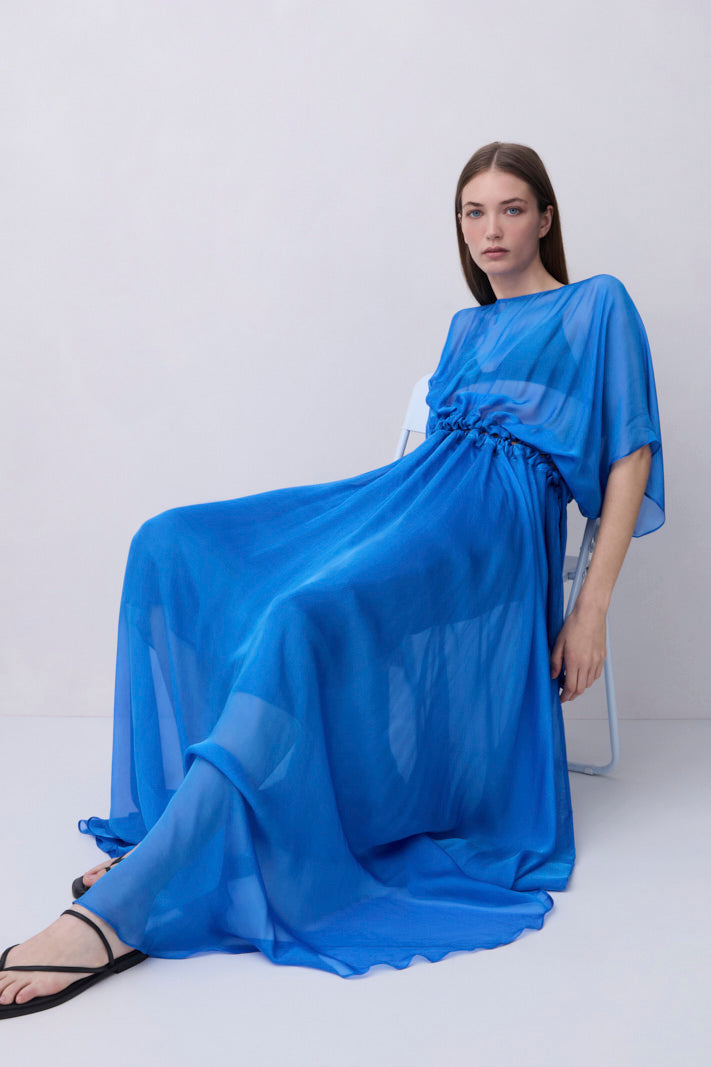 A woman wearing a flowing royal blue maxi dress with wide draped sleeves and a gathered waist sits on a white chair against a plain light gray studio background.
