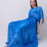 A woman wearing a flowing royal blue maxi dress with wide draped sleeves and a gathered waist sits on a white chair against a plain light gray studio background.