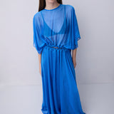 A woman modeling a sheer, flowing blue maxi dress with elbow-length sleeves and a gathered waist, set against a plain light gray studio background.
