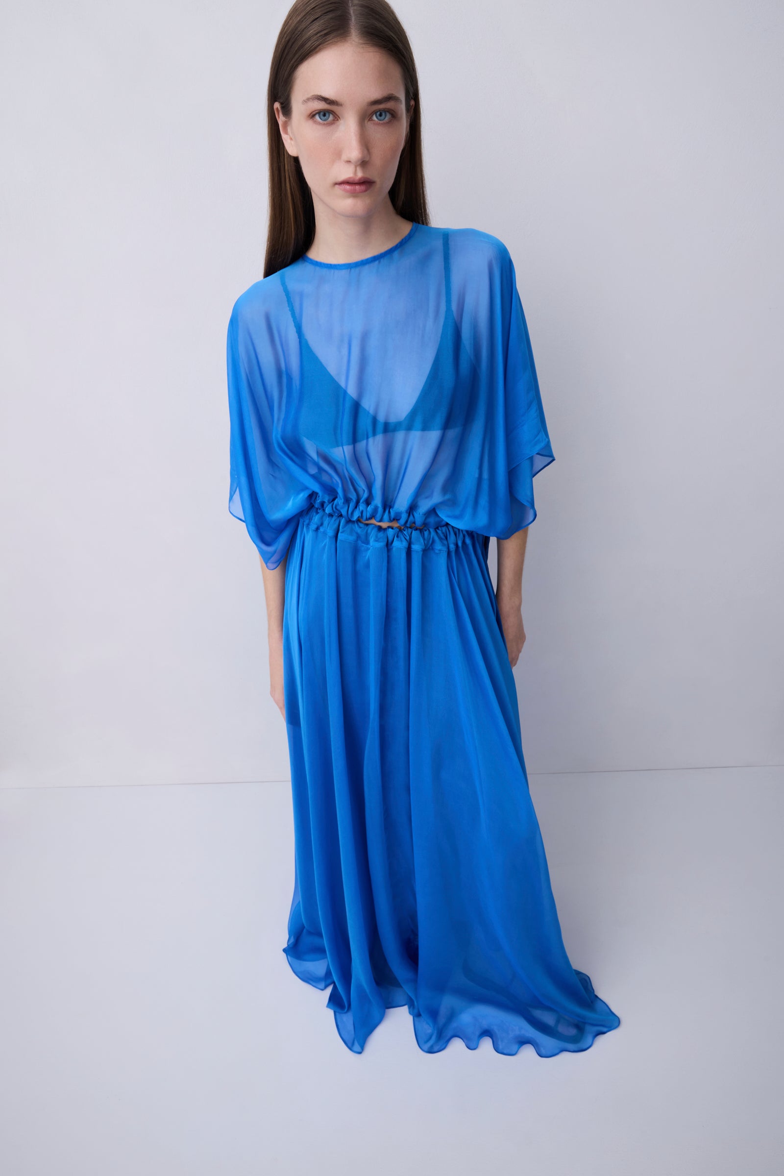 A woman modeling a sheer, flowing blue maxi dress with elbow-length sleeves and a gathered waist, set against a plain light gray studio background.