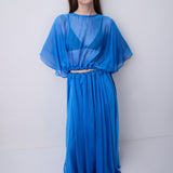 A woman modeling a vibrant blue sheer maxi dress with a draped, flowing silhouette, wide short sleeves, and a gathered waist detail, set against a plain light gray studio background.