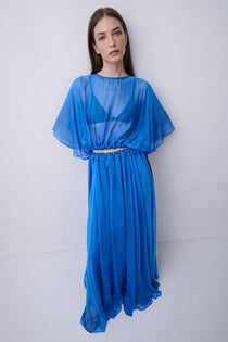 A woman modeling a vibrant blue sheer maxi dress with a draped, flowing silhouette, wide short sleeves, and a gathered waist detail, set against a plain light gray studio background.