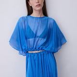 A woman modeling a vibrant blue sheer maxi dress with a draped, flowing silhouette, wide short sleeves, and a gathered waist detail, set against a plain light gray studio background.