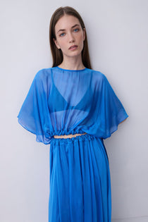 A woman modeling a vibrant blue sheer maxi dress with a draped, flowing silhouette, wide short sleeves, and a gathered waist detail, set against a plain light gray studio background.