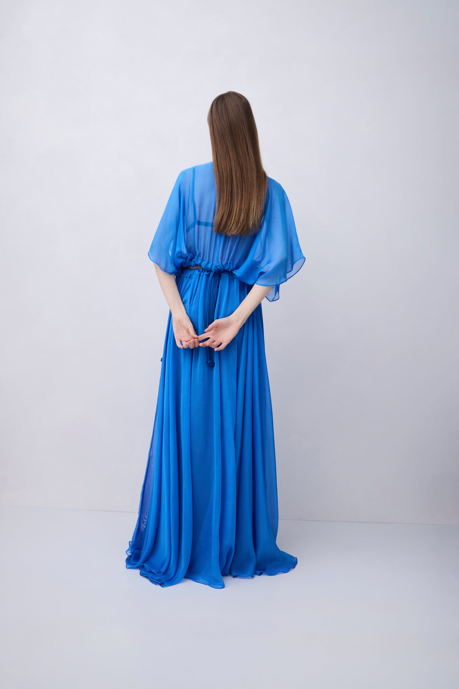 A woman with long brown hair is shown from behind wearing a flowing royal blue maxi dress with wide draped sleeves, standing against a plain light gray studio background.