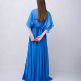 A woman with long brown hair is shown from behind wearing a flowing royal blue maxi dress with wide draped sleeves, standing against a plain light gray studio background.