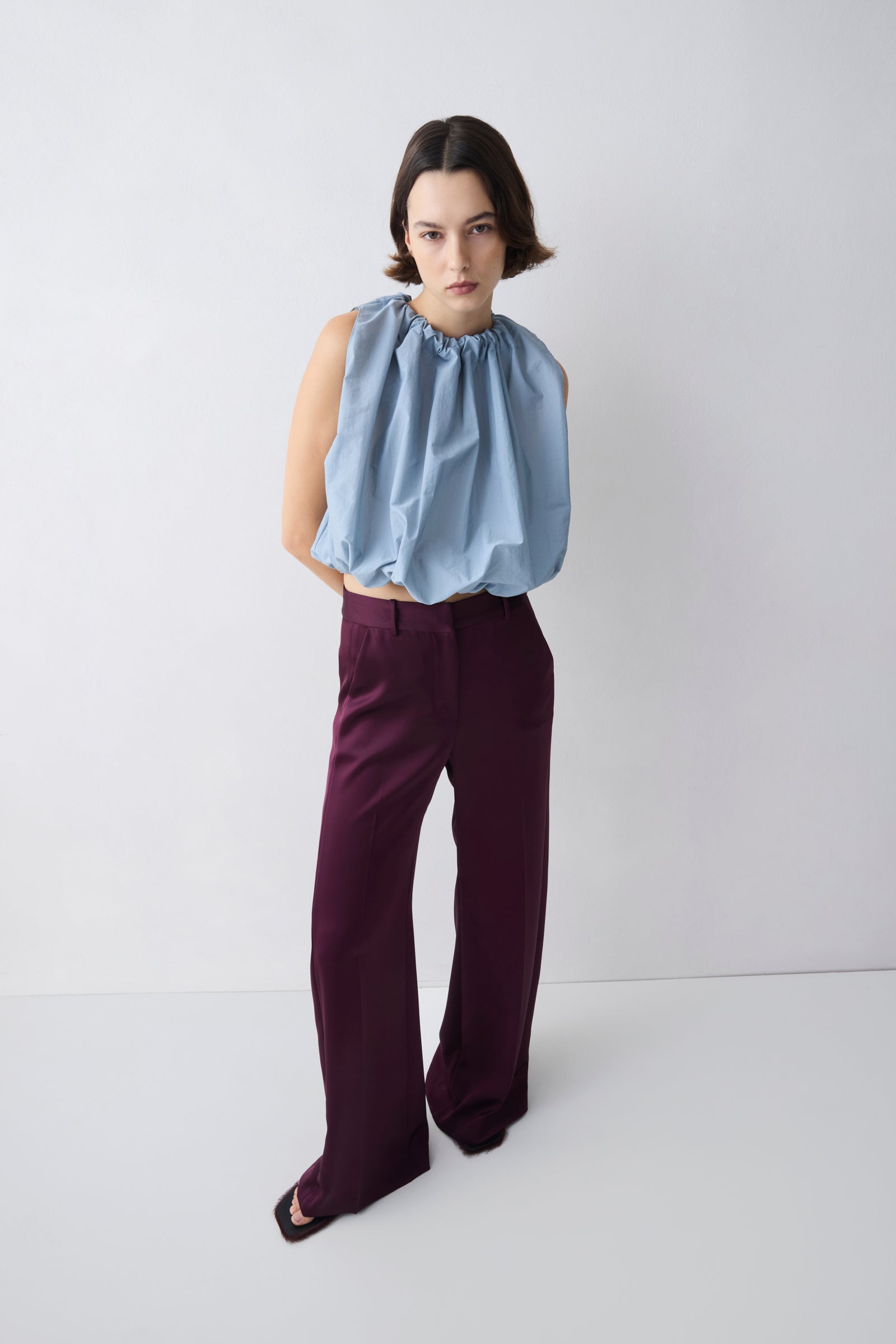 Woman wearing a light blue sleeveless top and dark purple pants on a white background