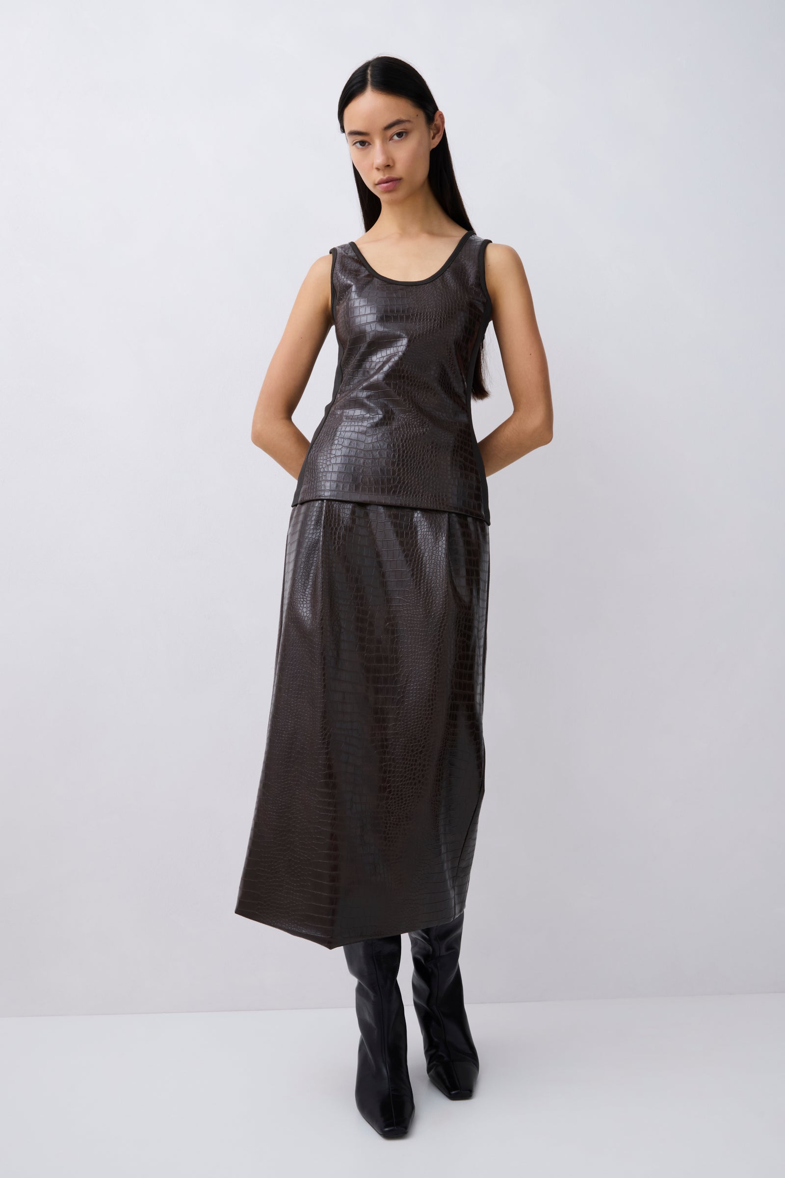 A model wearing a dark gray faux-leather sleeveless top and matching skirt set, paired with black boots, against a light gray studio background.