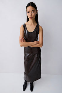 A model wearing a dark brown textured faux-leather sleeveless top and matching skirt set, paired with black pointed-toe boots, against a plain light gray studio background.