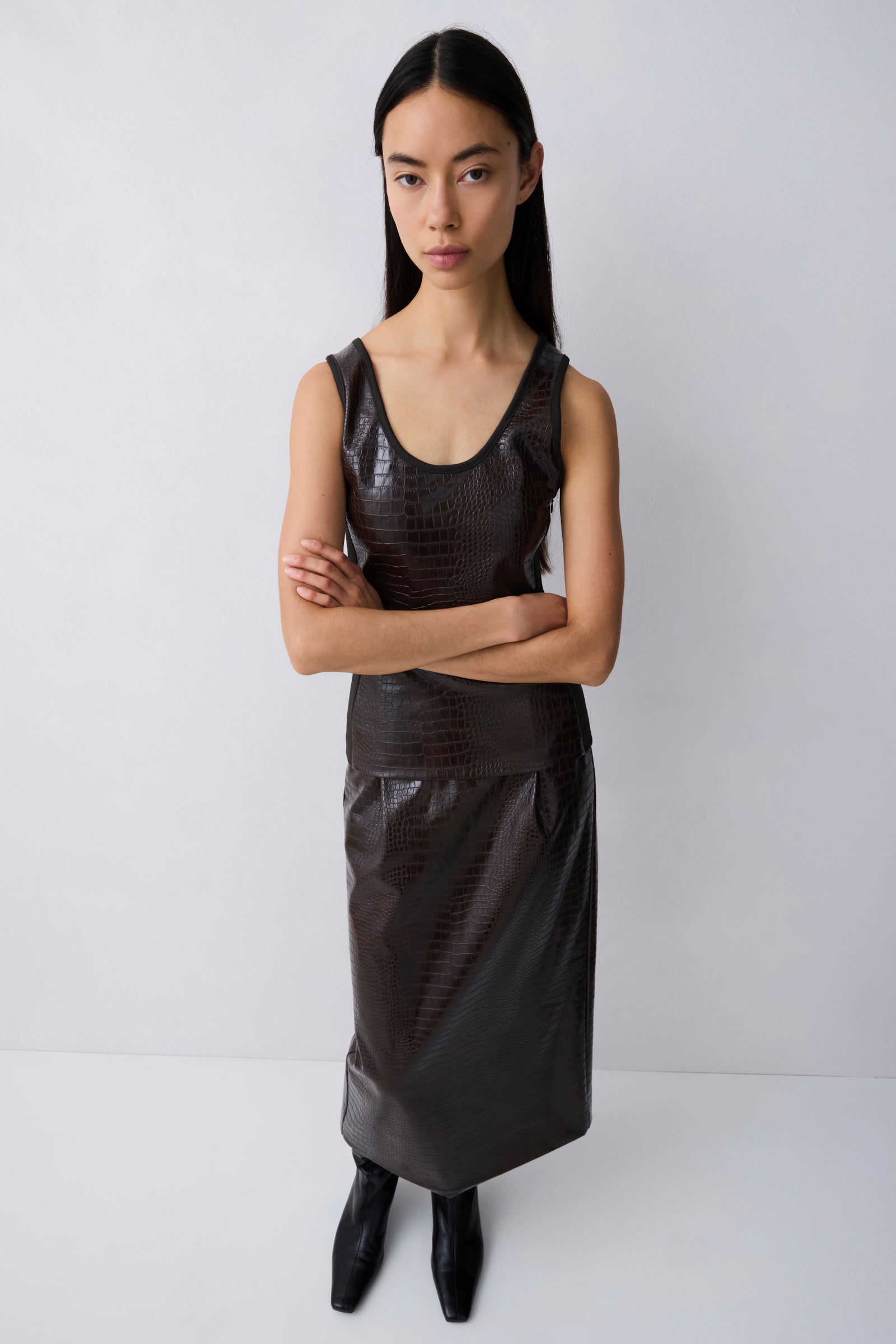 A model wearing a dark brown textured faux-leather sleeveless top and matching skirt set, paired with black pointed-toe boots, against a plain light gray studio background.