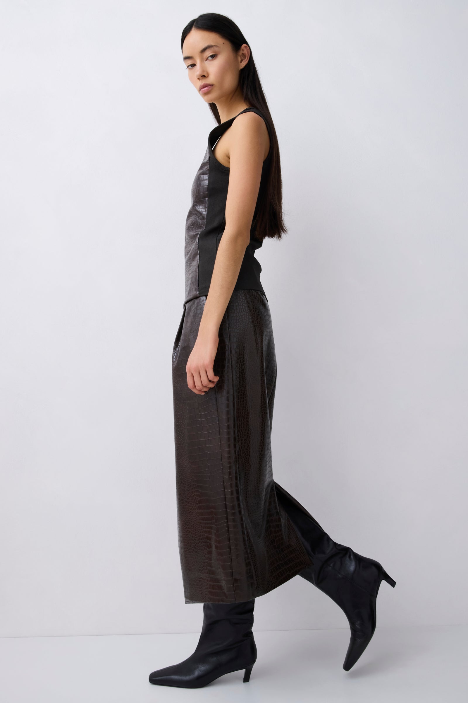 A model wearing a dark brown textured faux-leather sleeveless dress with wide legs and black pointed-toe heeled boots, shown in profile against a light gray studio background.