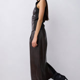 A model wearing a dark brown textured faux-leather sleeveless dress with wide legs and black pointed-toe heeled boots, shown in profile against a light gray studio background.
