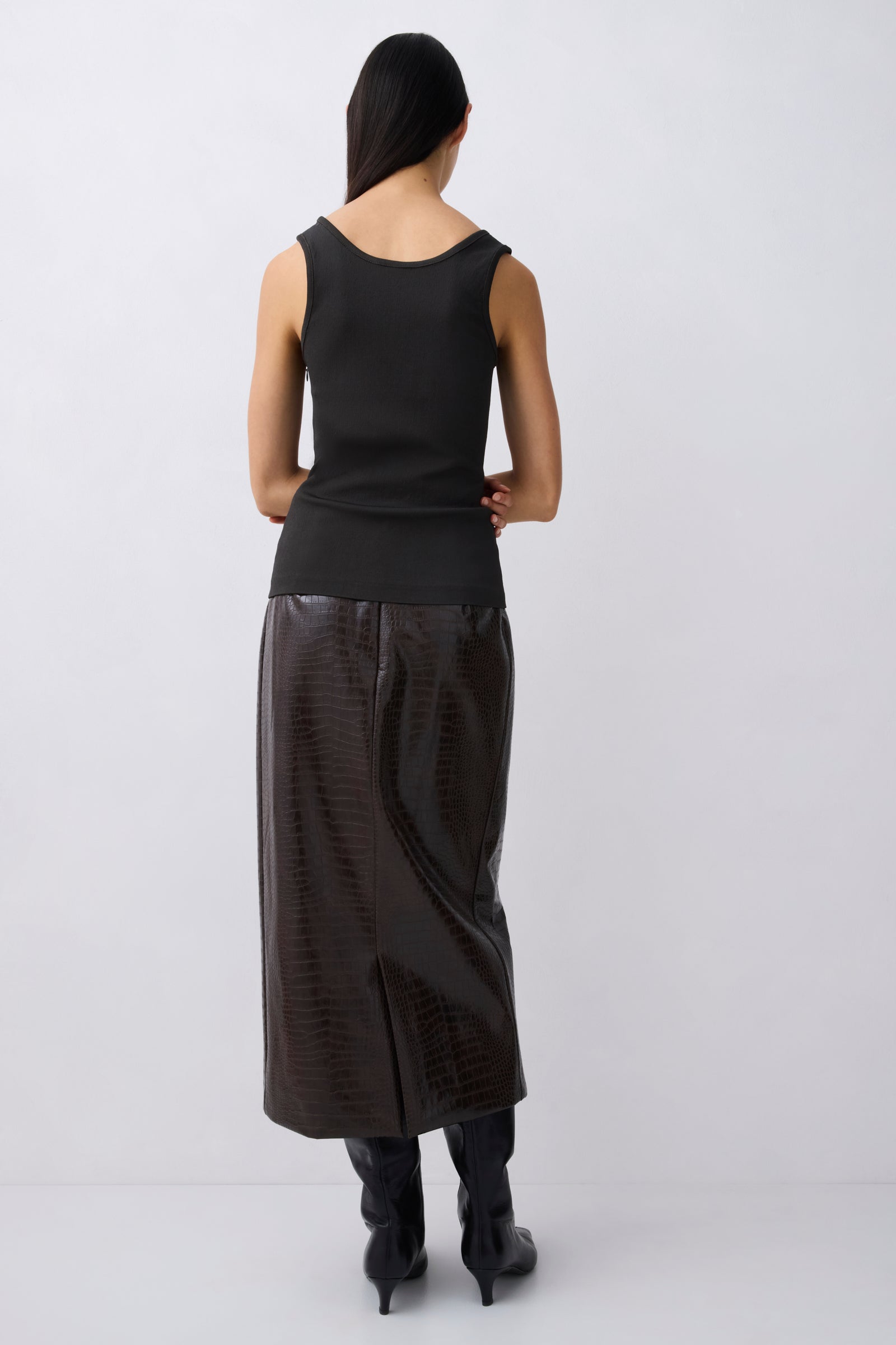 A model shown from the back wearing a black sleeveless top and a black textured midi skirt, paired with black heeled boots, against a light gray studio background.
