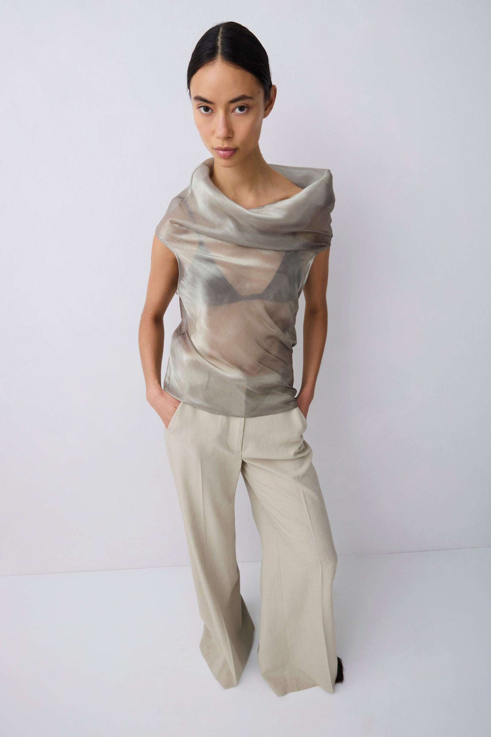 A model wearing a draped, semi-sheer metallic beige top paired with wide-leg beige trousers, standing against a plain light gray studio background.