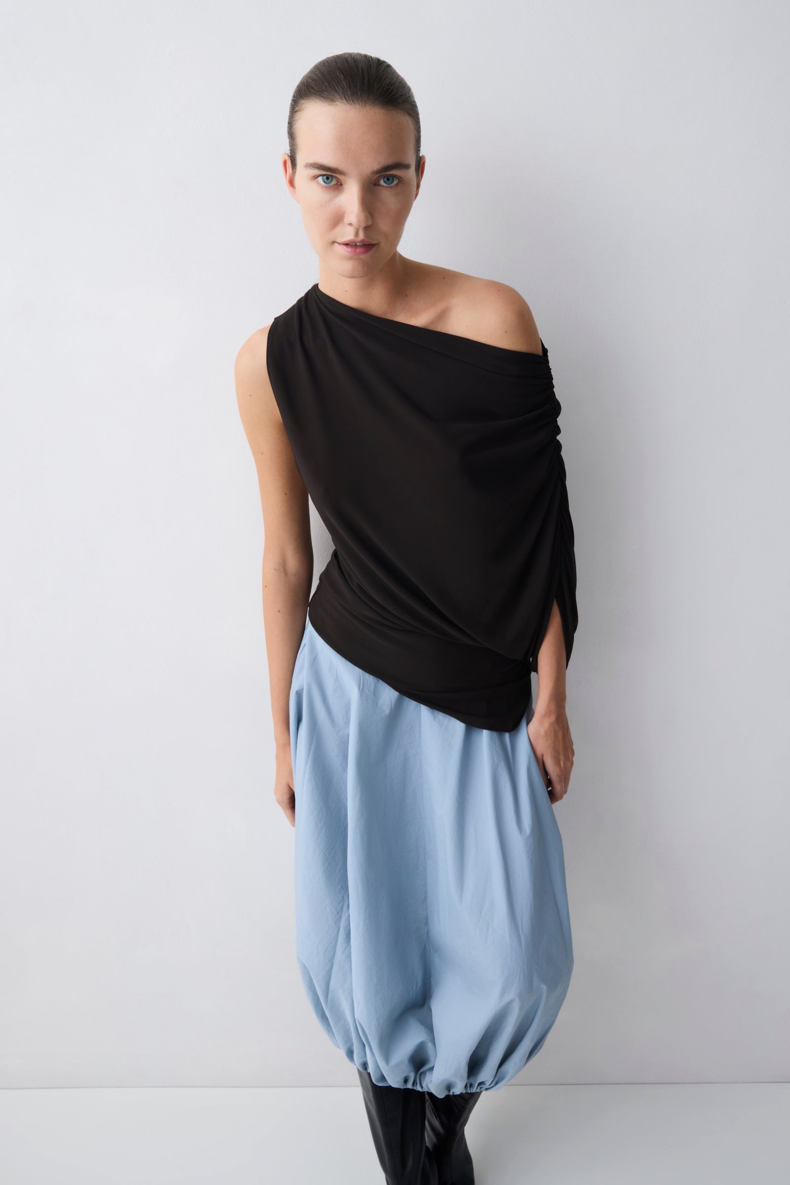 Woman wearing a brown one-shoulder top and light blue harem pants against a white background