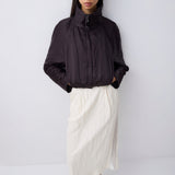 A model stands against a light gray studio background wearing a dark charcoal crinkled zip-up jacket with a high collar, paired with an off-white midi skirt and black pointed flats.
