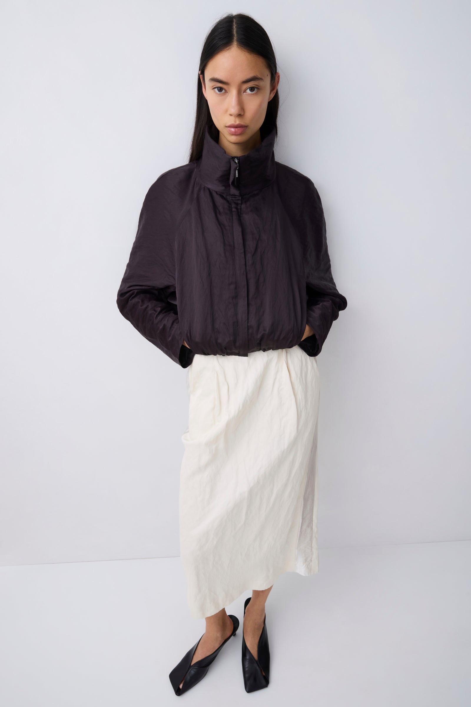 A model stands against a light gray studio background wearing a dark charcoal crinkled zip-up jacket with a high collar, paired with an off-white midi skirt and black pointed flats.