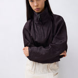 A model with long black hair is wearing a dark brownish-black lightweight zip-up windbreaker jacket with a high collar, paired with cream-colored trousers, against a plain light gray background.