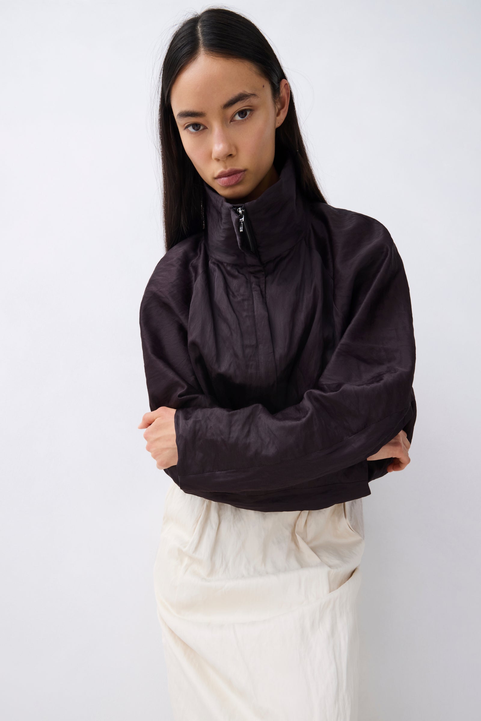 A model with long black hair is wearing a dark brownish-black lightweight zip-up windbreaker jacket with a high collar, paired with cream-colored trousers, against a plain light gray background.