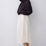 A woman stands in profile wearing a black cropped puffer jacket, an off-white midi skirt, and black pointed mule heels against a plain light gray studio background.
