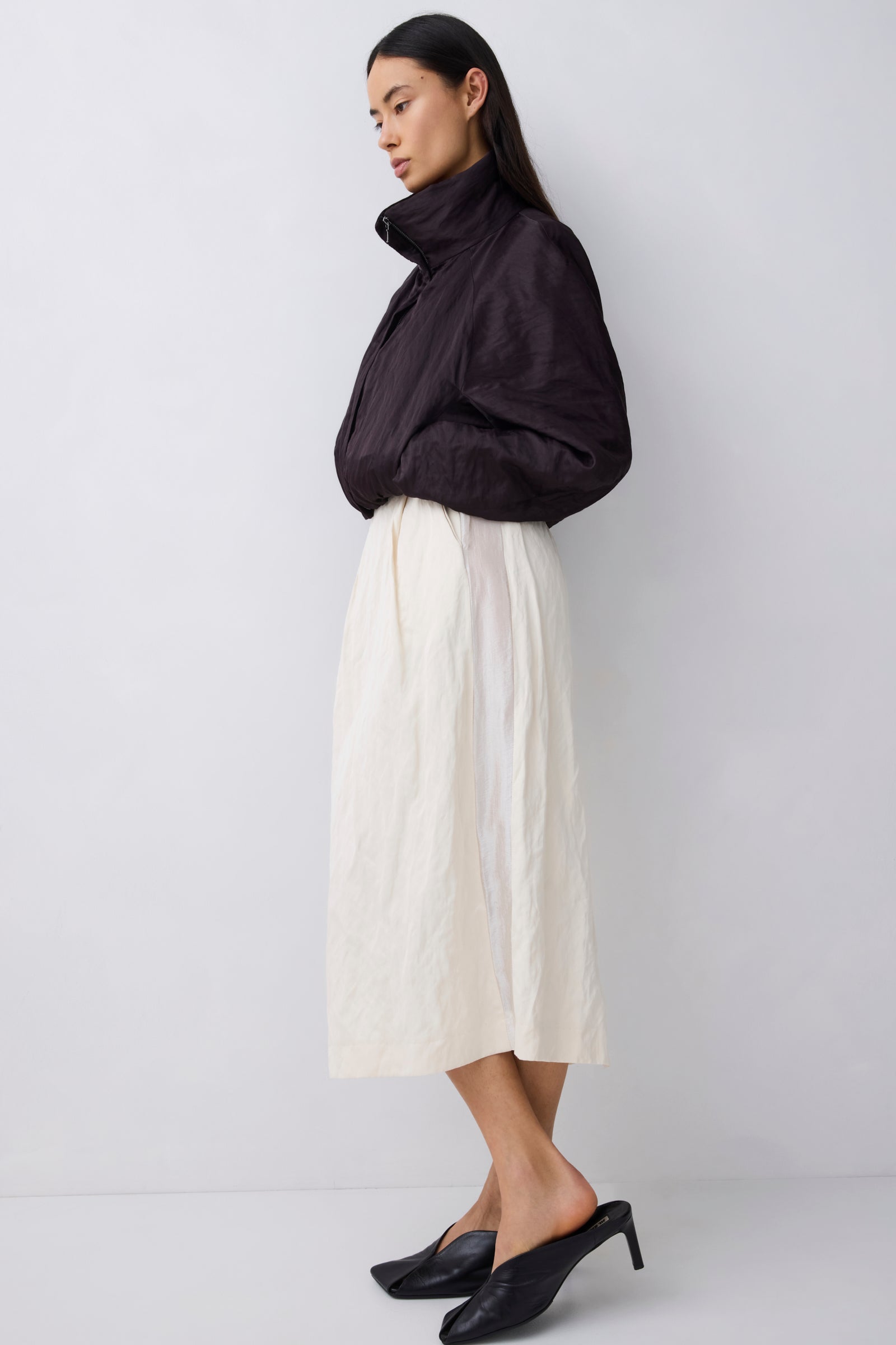 A woman stands in profile wearing a black cropped puffer jacket, an off-white midi skirt, and black pointed mule heels against a plain light gray studio background.