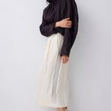 A model wearing a black high-neck long-sleeve top and a cream pleated midi skirt, paired with black pointed-toe mule heels, standing against a light gray studio background.