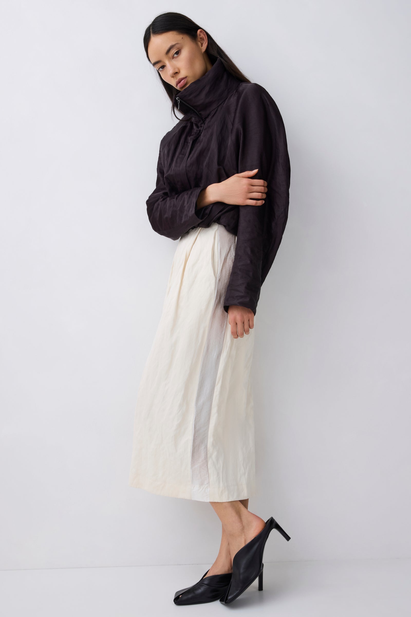 A model wearing a black high-neck long-sleeve top and a cream pleated midi skirt, paired with black pointed-toe mule heels, standing against a light gray studio background.