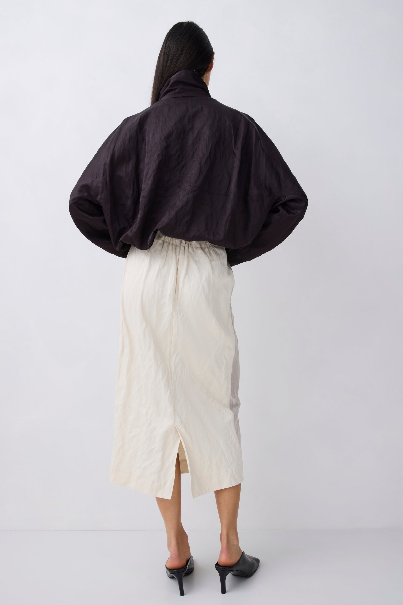 A model shown from the back wearing a black cropped, voluminous jacket and an off-white midi skirt, paired with black pointed-toe mule heels, against a light gray studio background.