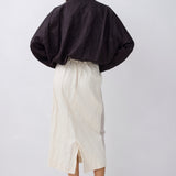 A model shown from the back wearing a black cropped, voluminous jacket and an off-white midi skirt, paired with black pointed-toe mule heels, against a light gray studio background.
