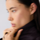 A close-up side profile of a woman with dark hair wearing a dark, high-collar jacket against a plain light background.