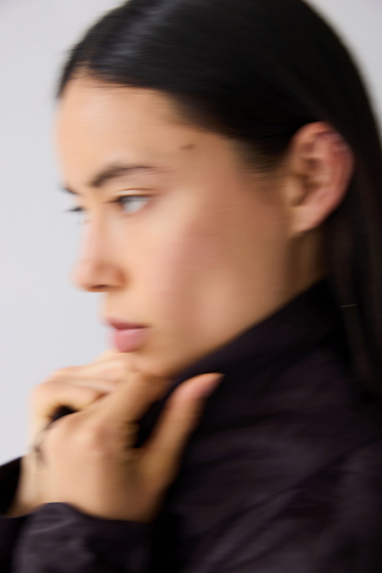 A close-up side profile of a woman with dark hair wearing a dark, high-collar jacket against a plain light background.
