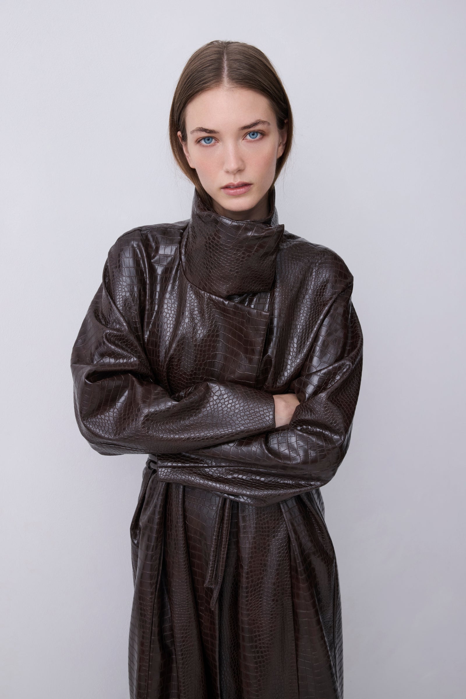 A model wearing a dark brown textured faux-leather wrap coat with a high collar and long sleeves, standing against a light gray background.